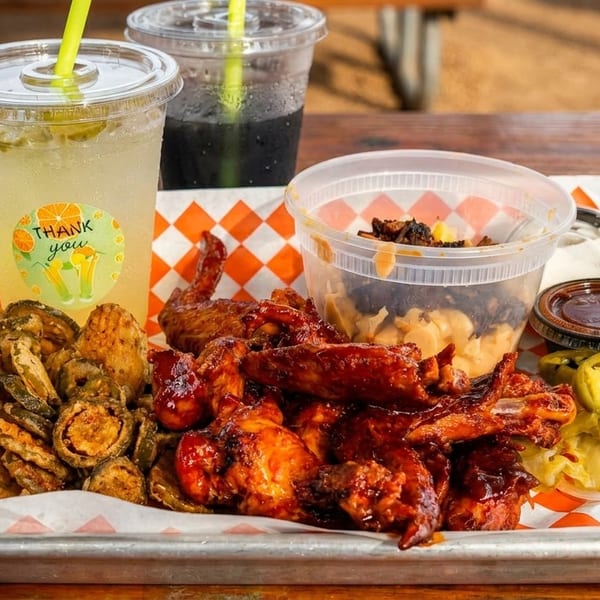 A metal tray with BBQ wings, fried pickles, mac and cheese, pickled jalapeños, and two drinks.