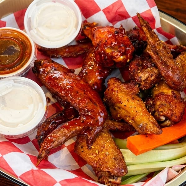 A platter of chicken wings with two dipping sauces, carrots, and celery on a red and white checkered paper.