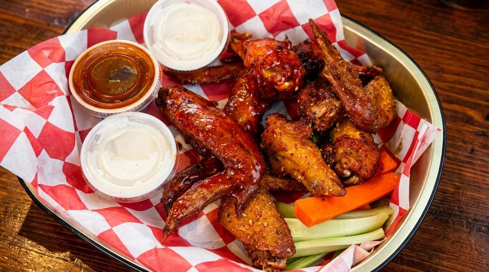 A platter of chicken wings with two dipping sauces, carrots, and celery on a red and white checkered paper.