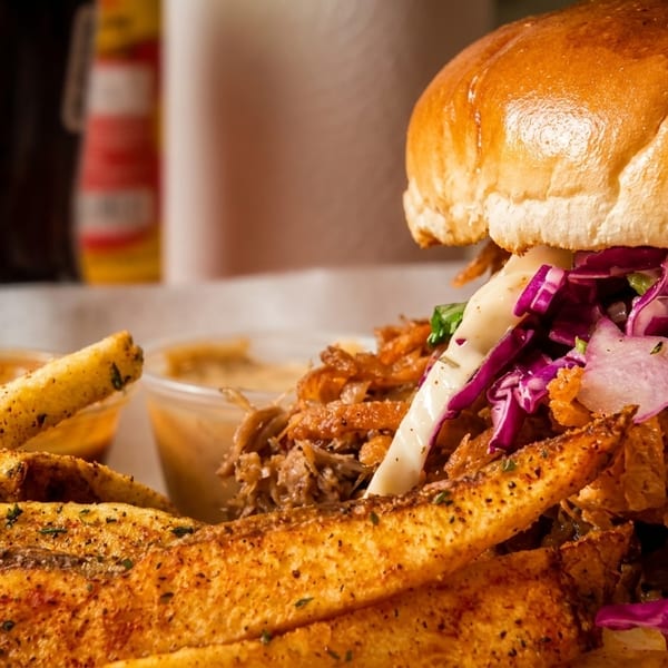 Close-up of a pulled pork sandwich with purple coleslaw and a side of seasoned fries.