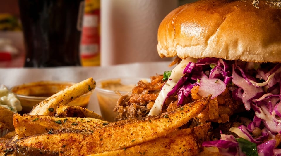 Close-up of a pulled pork sandwich with purple coleslaw and a side of seasoned fries.